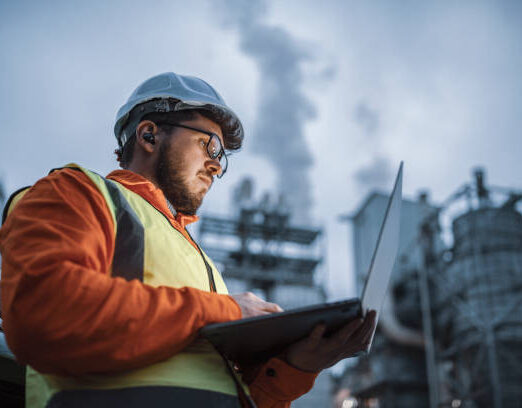 A shot of an young engineer wearing a helmet and using a laptop and hands free device during his night shirt in the oil rafinery. Engineering concept.